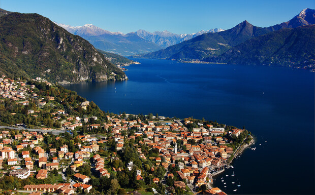 A picturesque town with red-roofed buildings lines a serene lakeshore, surrounded by mountains and lush greenery under a clear blue sky. Snow-capped peaks are visible in the distance.